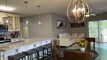 A modern kitchen with a marble countertop, bar stools, a dining table with chairs, and a chandelier.