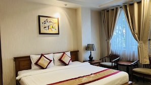 Quarto de hotel com cama, cadeira, mesa pequena, abajur e janela com cortinas.
