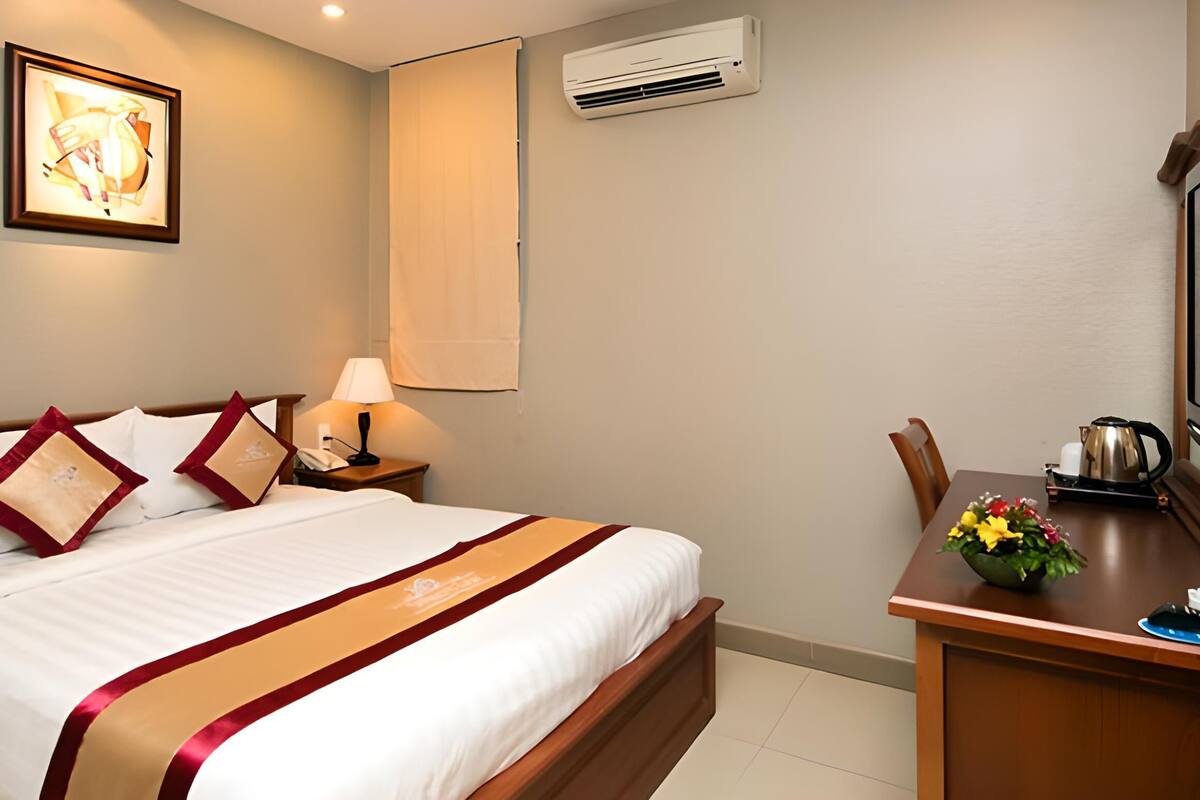 Habitación de hotel con cama, mesita de noche, lámpara, equipo de aire acondicionado, cuadro enmarco y escritorio con hervidor y flores.
