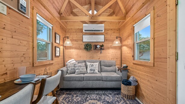 A cozy cabin interior with a wooden ceiling, a gray sofa with cushions, a dining table with chairs, and a large window. - Lolli's Lil Getaway (Alma)