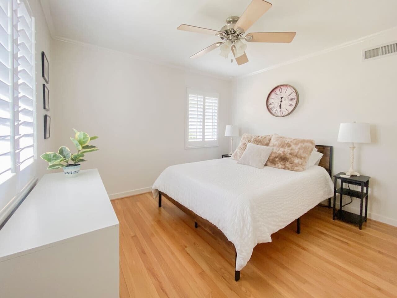 A bedroom with a bed, bedside tables, a ceiling fan, a clock, and a window with blinds.
