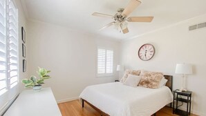 A bedroom with a bed, bedside tables, a ceiling fan, a clock, and a window with blinds.
