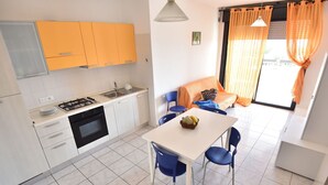 A compact kitchen with white and yellow cabinets, a gas stove, and a sink. There is a dining table with blue chairs and a bowl of fruit. Orange curtains are visible in the background. - Nice apartment for 4 guests with WIFI, TV, balcony and pets allowed (Silvi)
