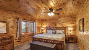 A wooden cabin bedroom with a bed, nightstands, a bench, a ceiling fan, and a window with curtains.