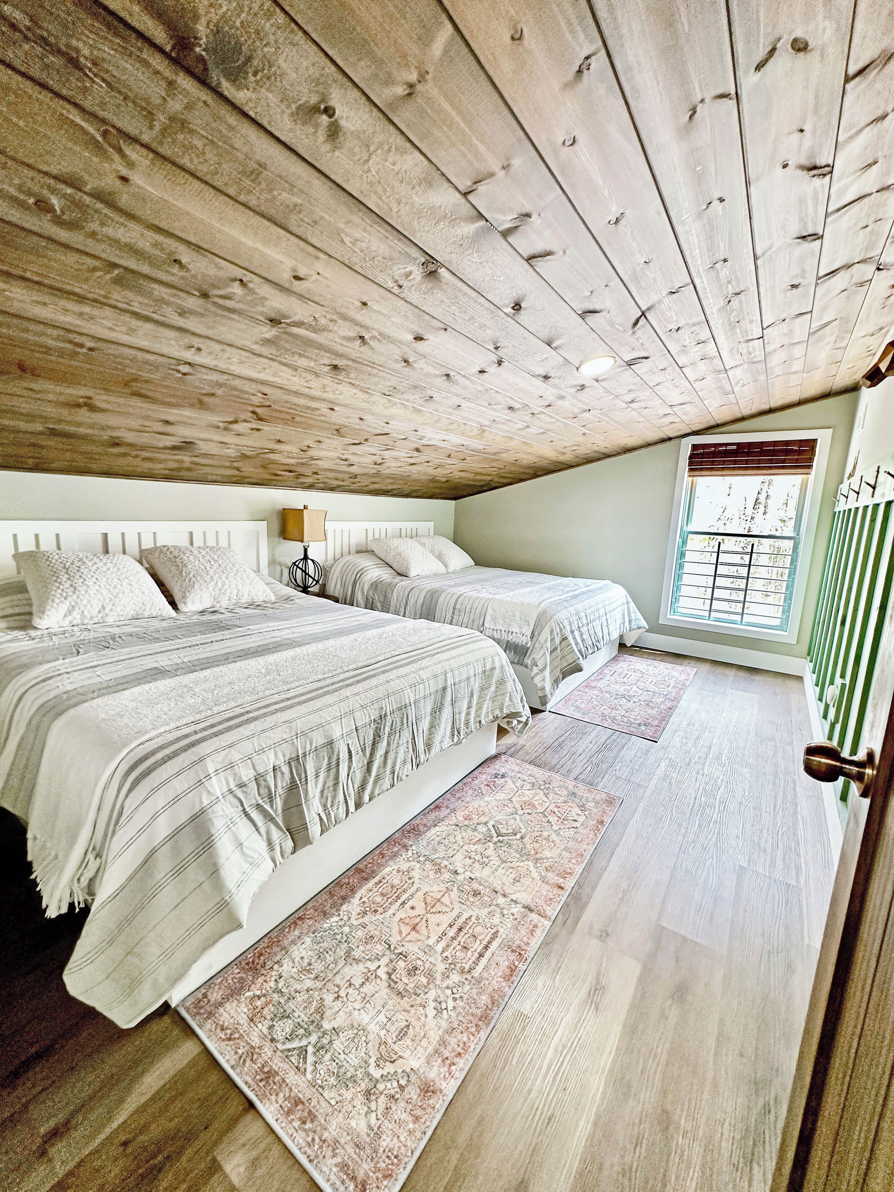 A bedroom with two beds, a window with blinds, a patterned rug, and a wooden ceiling.