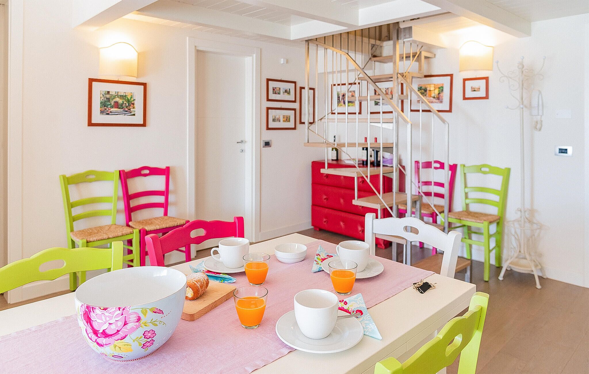 A dining area with a table set for breakfast, chairs, a staircase, and framed pictures on the walls.