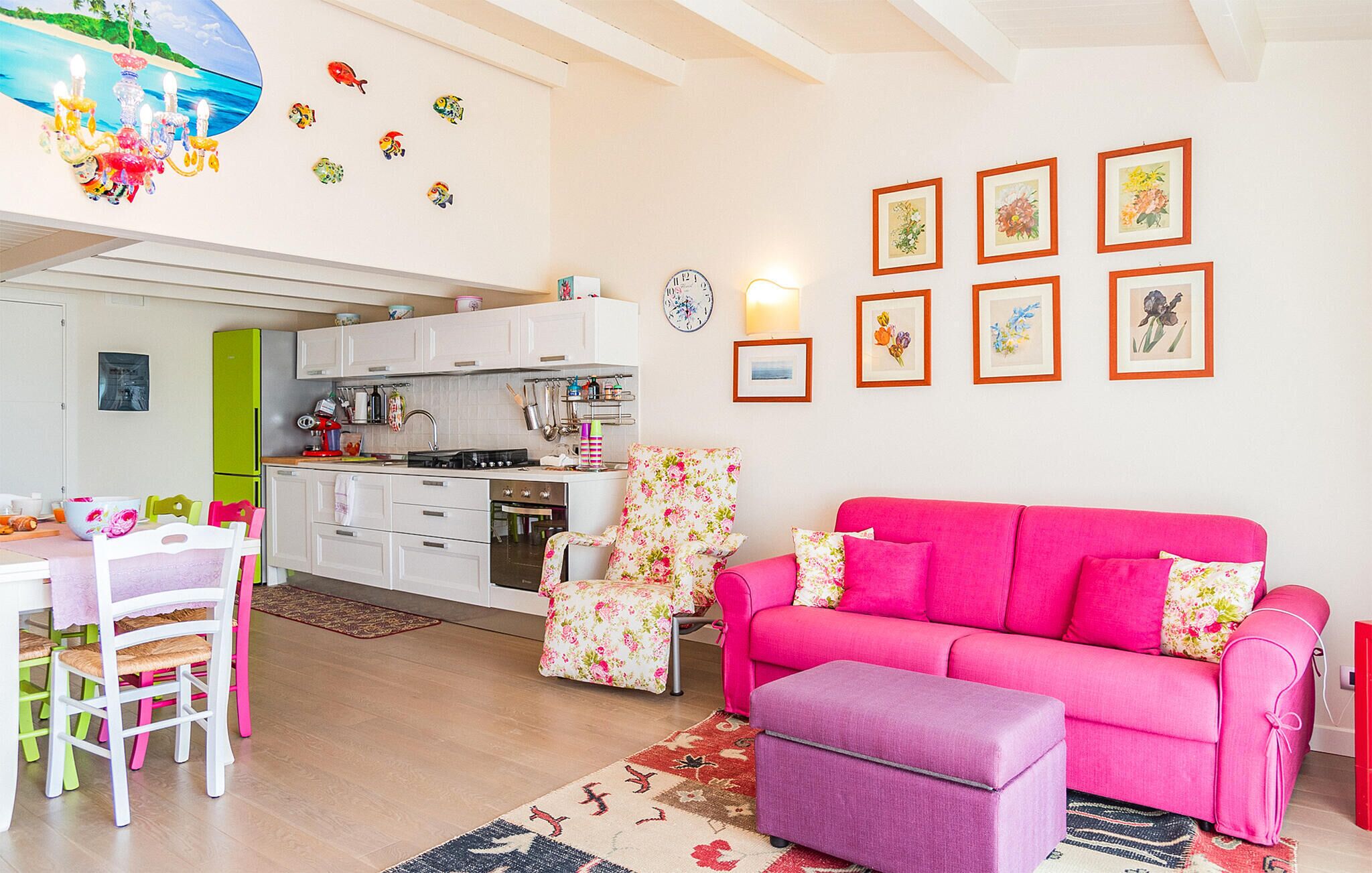 Un salón luminoso con un sofá rosa, un reposapiés púrpura y una zona de cocina con armarios blancos y un refrigerador verde.