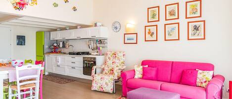 Un salón luminoso con un sofá rosa, un reposapiés púrpura y una zona de cocina con armarios blancos y un refrigerador verde.