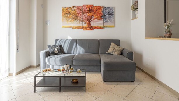 Ein modernes Wohnzimmer mit einem grauen Ecksofa, einem Glastisch und einem großen Wandbild mit Herbstmotiven.