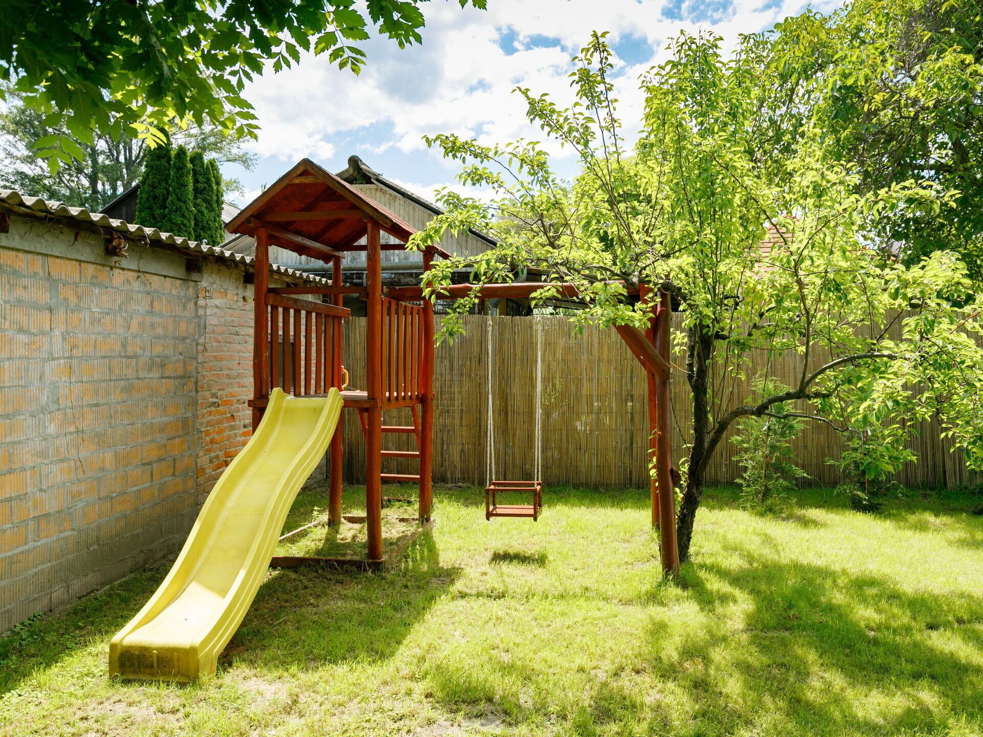 A wooden play structure with a slide and swing, surrounded by a grassy yard and a wooden fence.