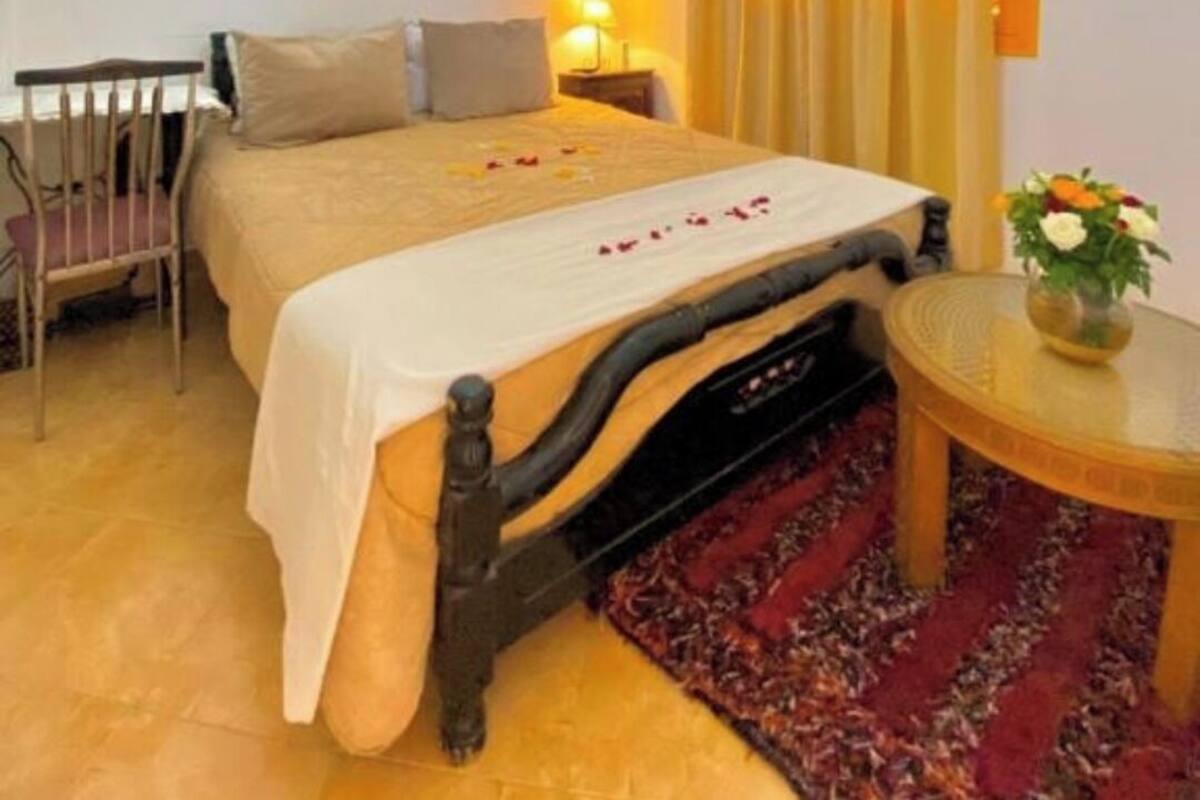 Une chambre d’hôtel avec un lit, une chaise, une petite table avec un vase et une fenêtre avec des rideaux.