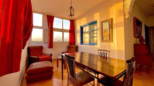 Uma sala de jantar com uma longa mesa de madeira, cortinas vermelhas e vista para a cidade pelas janelas.