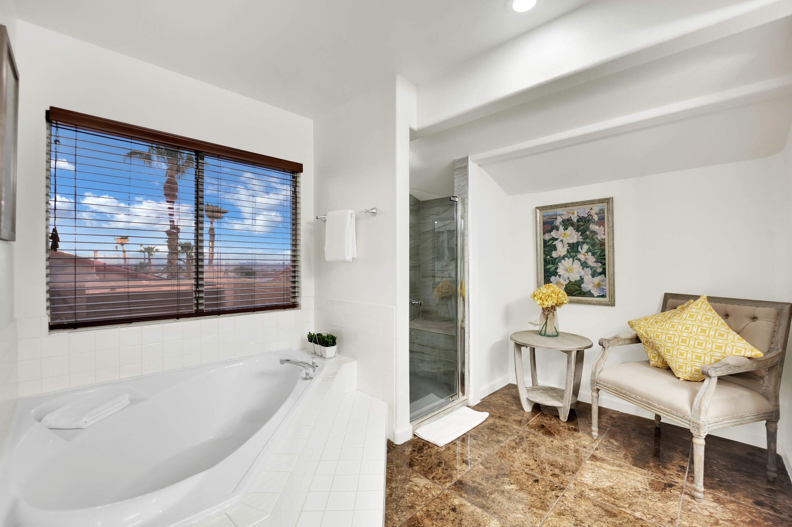A bathroom with a bathtub, a glass shower enclosure, a small round table with a vase, and a cushioned chair with a patterned pillow.