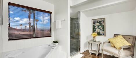 A bathroom with a bathtub, a glass shower enclosure, a small round table with a vase, and a cushioned chair with a patterned pillow.