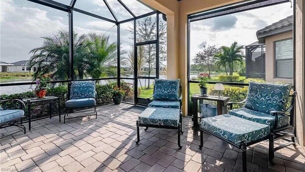 A covered patio with two cushioned chairs and a small table, overlooking a landscaped area with palm trees and a view of a house. - Welcome to this Endearing Naples Home! (Naples)