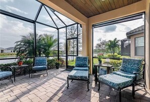 Une terrasse couverte avec deux fauteuils confortables et une petite table, donnant sur un espace aménagé avec des palmiers et offrant une vue sur une maison.