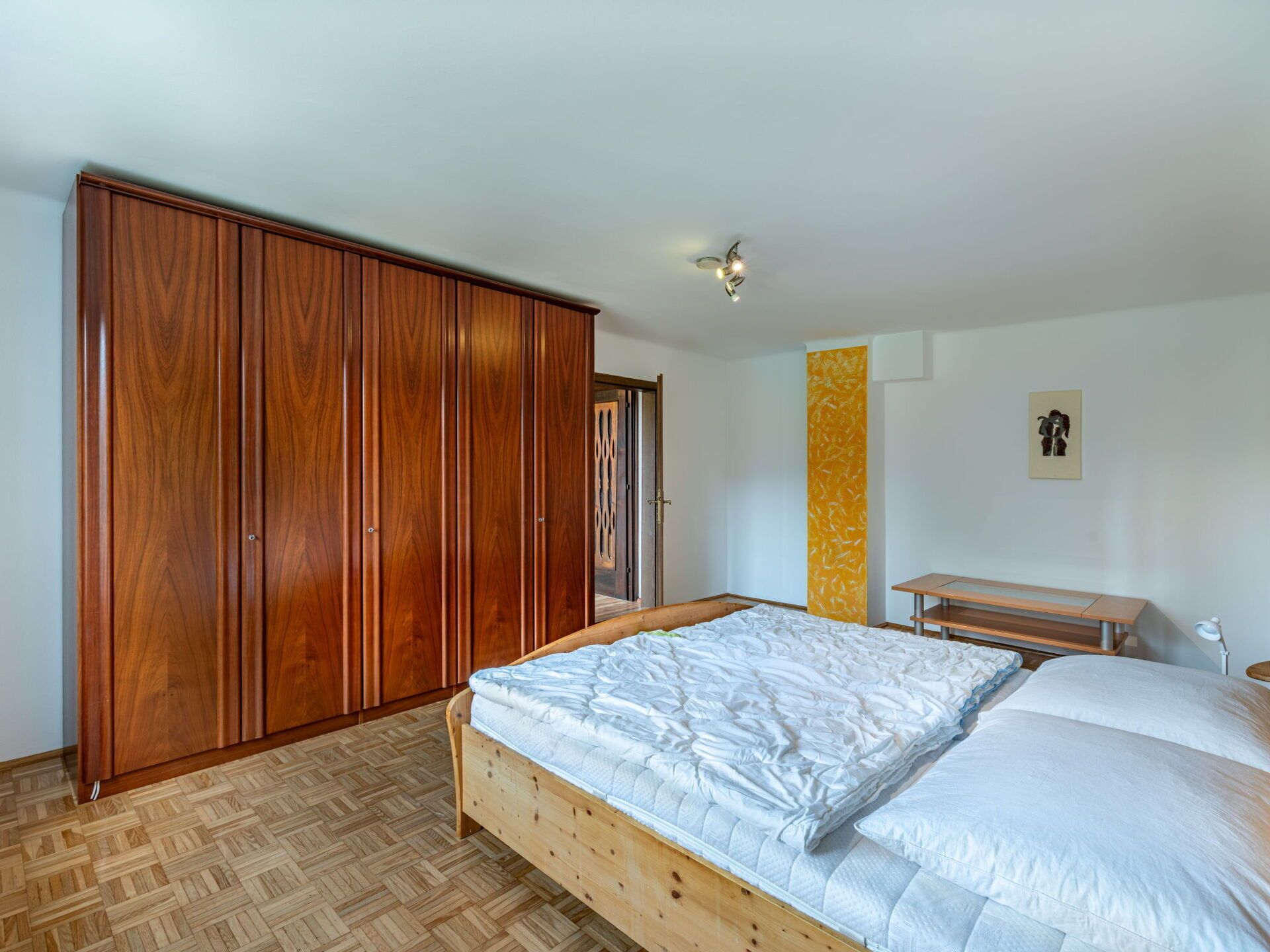 Un dormitorio con un armario de madera, una cama con ropa de cama blanca, una mesita pequeña y una pared con un diseño abstracto en color amarillo.