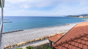 A coastal view with a pebbled beach, a red-roofed building, and a distant cityscape. - Lovely apartment for 4 guests with WIFI, TV and balcony (Riva Ligure)