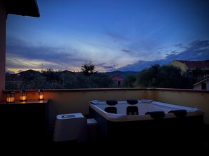 A balcony with a hot tub, a table with candles, and a view of houses and trees under a twilight sky.