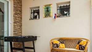 A room with a foosball table, a wicker sofa with patterned cushions, and two windows with decorative items.