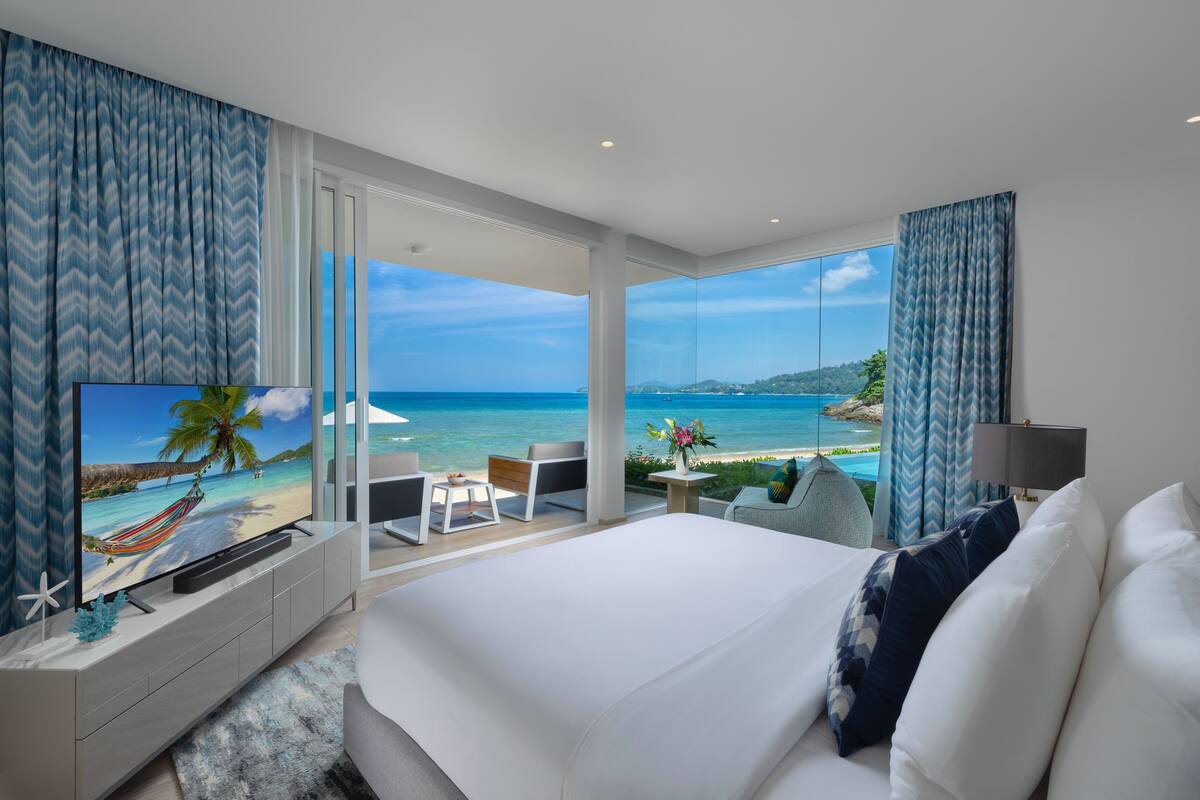 Een moderne hotelkamer met een groot bed, een televisie waarop een strandscène te zien is, een bureau en uitzicht op de oceaan.