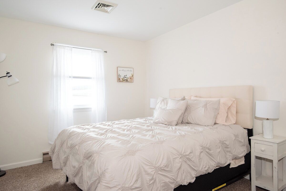 A bedroom with a bed, a nightstand, a lamp, a window with curtains, and a wall-mounted picture.