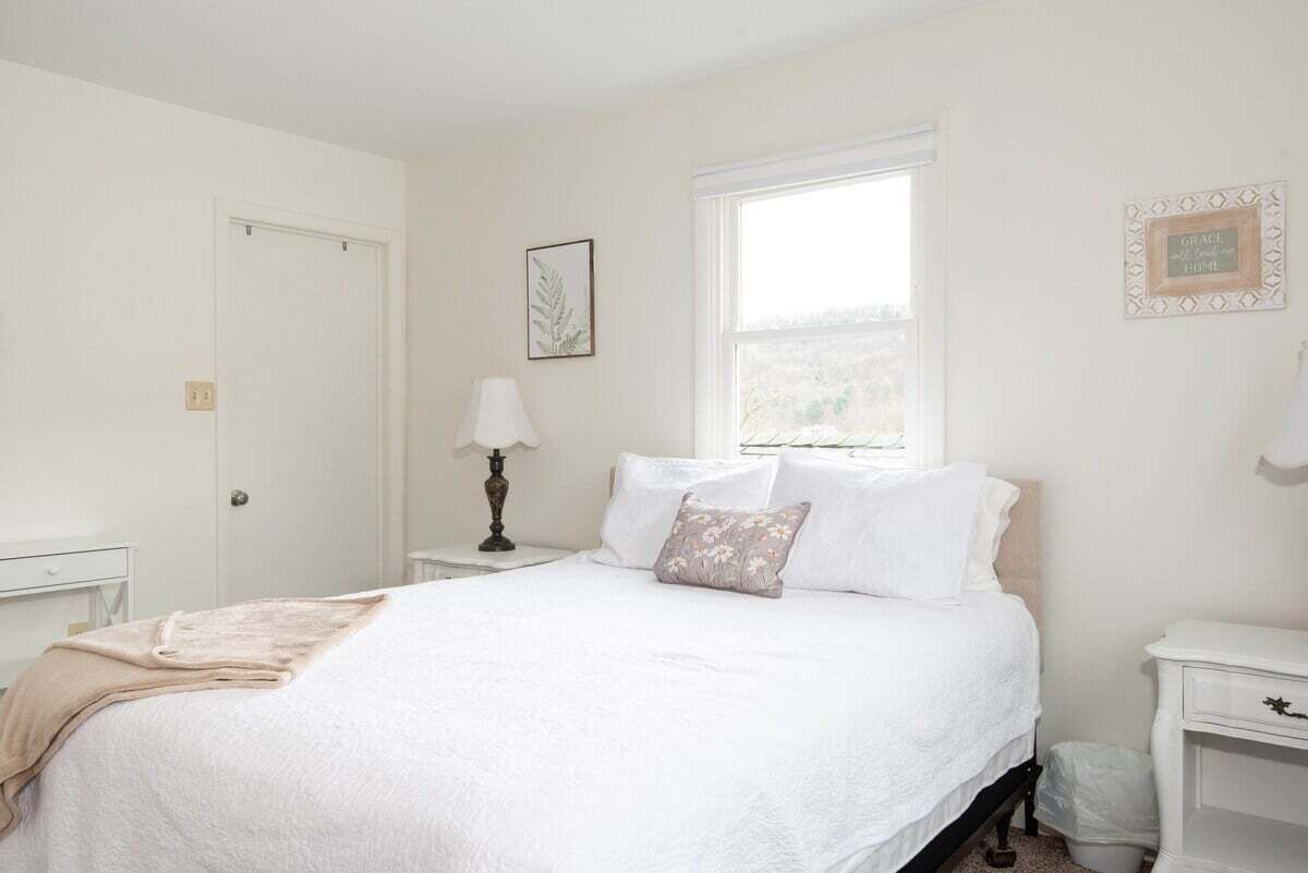 A bedroom with a bed, a bedside table, a lamp, a window, and a door.