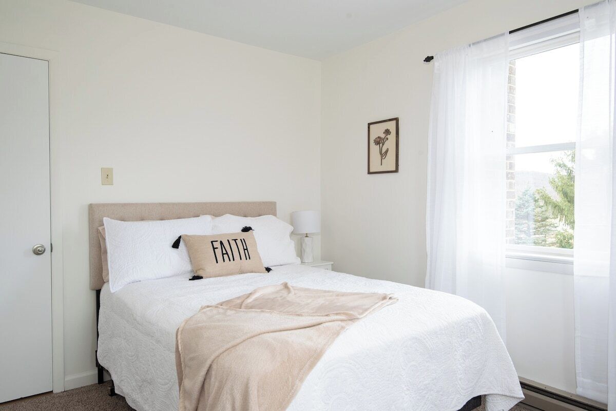 A bedroom with a bed, a bedside lamp, a window with curtains, and a wall-mounted picture.