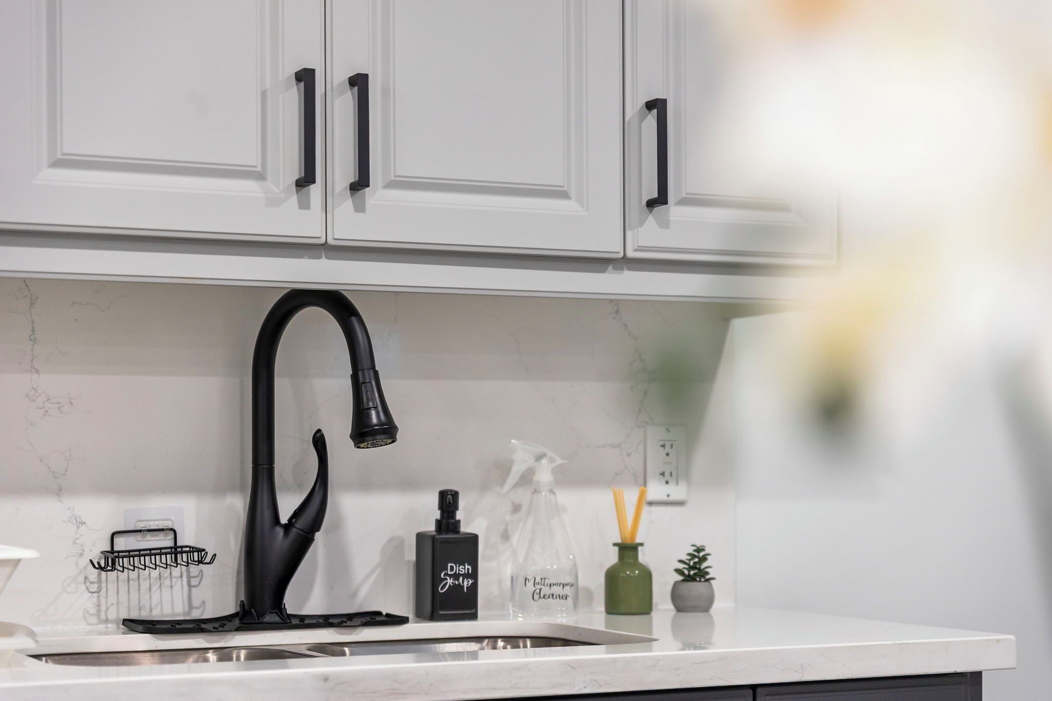 A kitchen with white cabinets, a black faucet, a dish soap bottle, a spray cleaner bottle, and a small green vase with a plant.