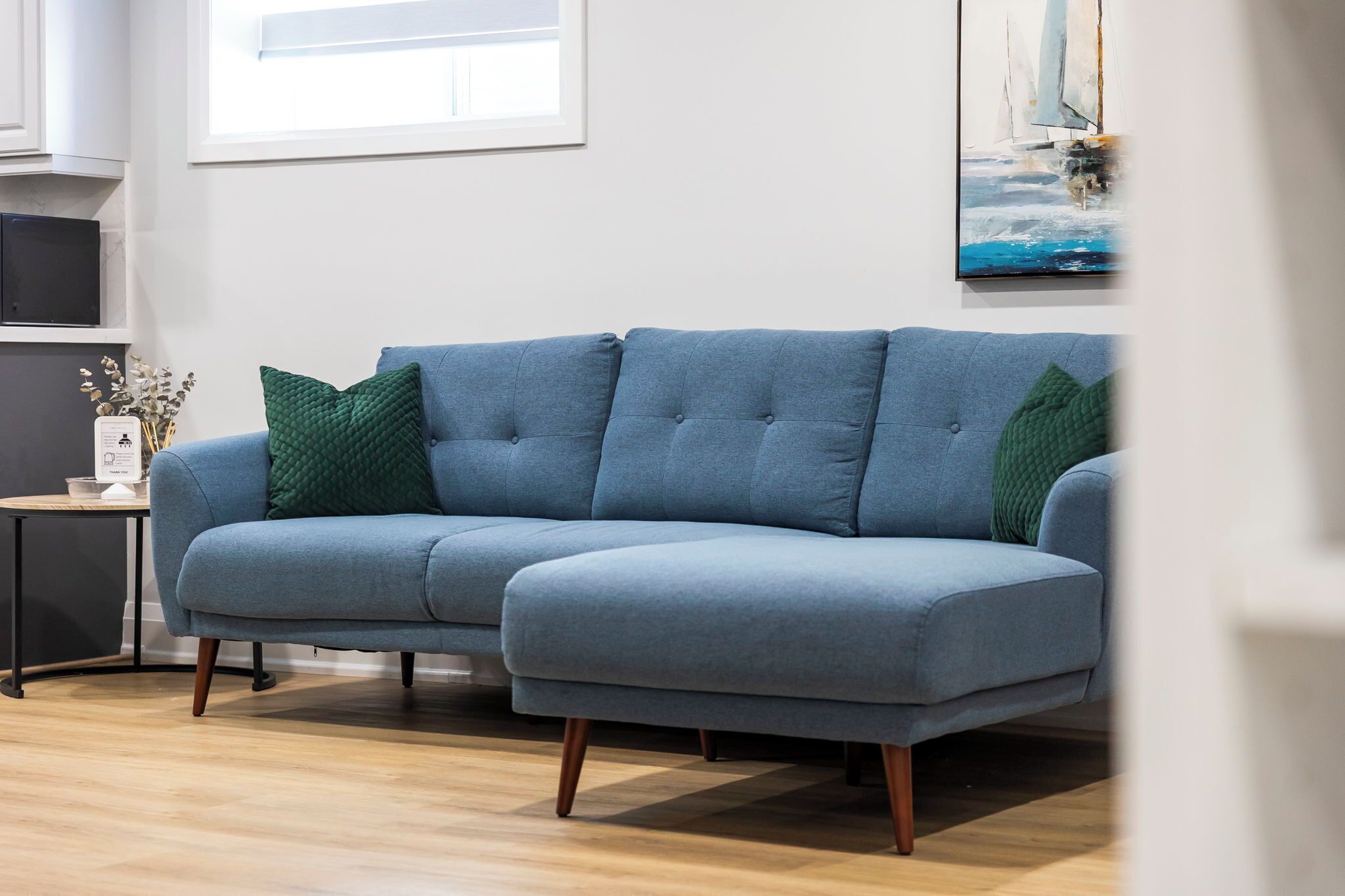 A light blue sectional sofa with green cushions, a small side table with a vase, and a painting on the wall.