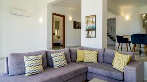 A modern living room with a grey sectional sofa, a dining area with a table and chairs, and a staircase leading to another level. - Joy of the Sea Villa (Agios Nikolaos)