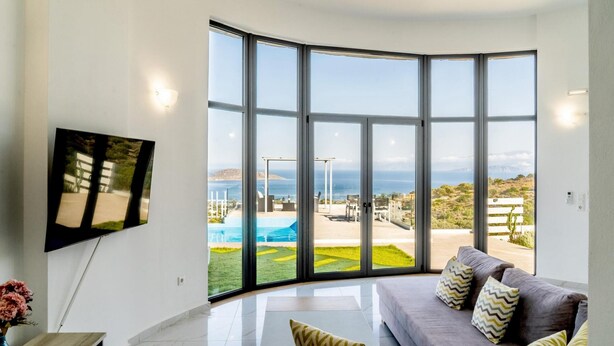 A modern living room with a large sliding glass door leading to a pool area, a flat-screen TV mounted on the wall, and a grey sofa with patterned cushions. - Joy of the Sea Villa (Agios Nikolaos)