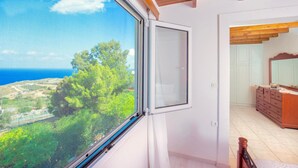 Quarto com uma grande janela que oferece vista para o mar e vegetação.
