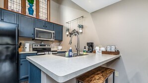 Een keuken met donkerblauwe kastjes, een wit aanrecht, een roestvrijstalen spoelbak, een magnetron, een koffiezetapparaat en een houten kruk.