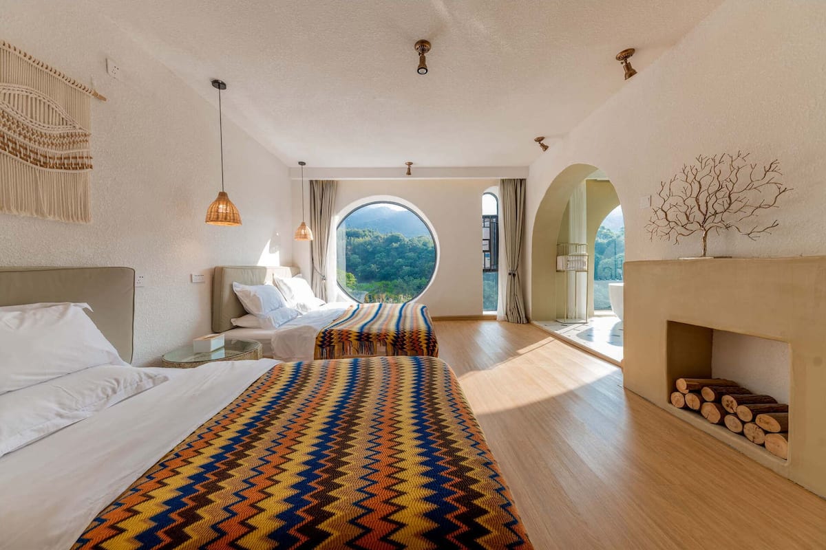 Ein geräumiges Schlafzimmer mit einem großen Bett, Blick ins Freie durch ein rundes Fenster und einem Kamin mit Holzscheiten.
