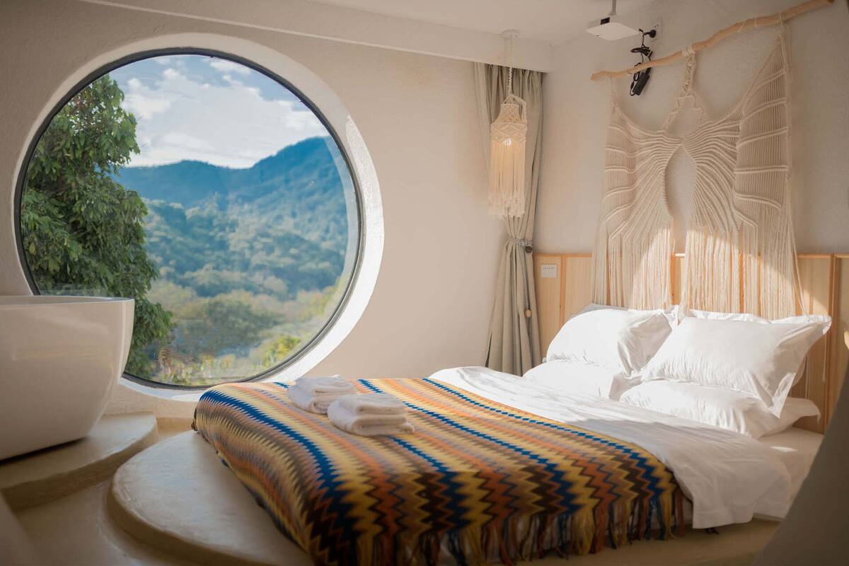 Ein Schlafzimmer mit einem großen Rundfenster, einem Bett mit weißer Bettwäsche, einer farbenfrohen Webdecke und einer Wandbehang aus Makramee.