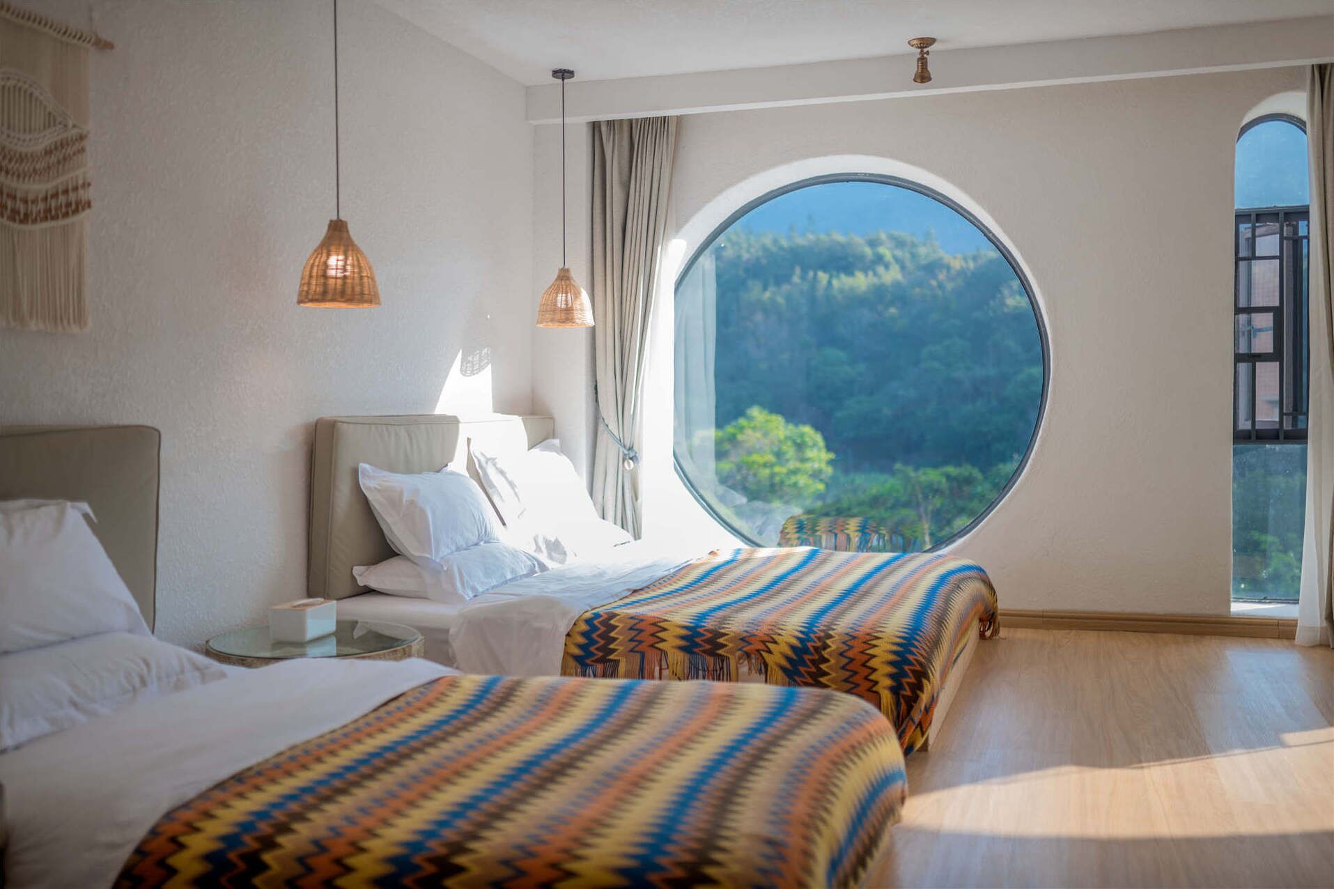 Un dormitorio con dos camas, un ventanal redondo y vistas al verdor exterior.