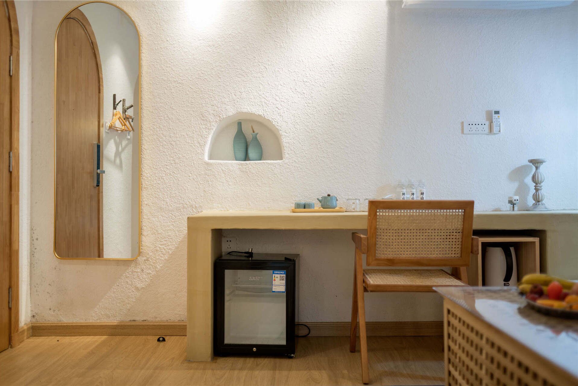 Une pièce de taille modeste, comprenant un bureau en bois, un petit réfrigérateur, une chaise et une étagère ornée d’objets décoratifs.