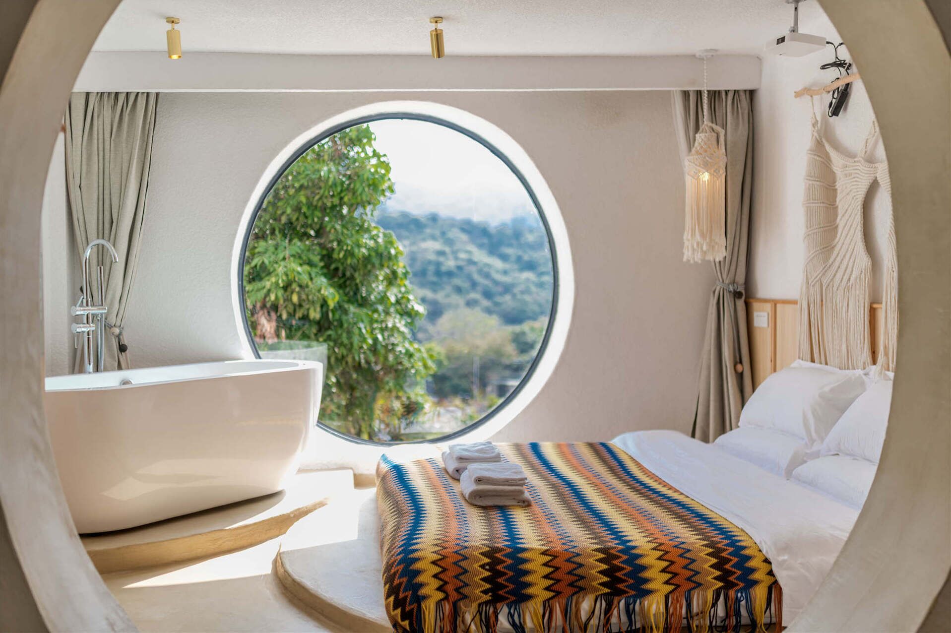 A bedroom with a large round window, a bathtub, a bed with a colorful blanket, and a macramé wall hanging.