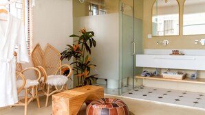 Un baño con cabina de ducha de vidrio, un lavabo blanco y un espejo. Hay sillas de mimbre, un banco de madera y un otoman redondo con estampado a rayas.