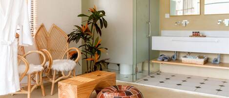 A bathroom with a glass shower enclosure, a white sink, and a mirror. There are wicker chairs, a wooden bench, and a round ottoman with a striped pattern.