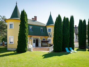 Un edificio grande de varios pisos con techo verde y múltiples torres. Cuenta con un césped bien cuidado con dos sillones azules.