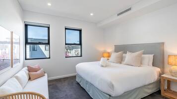 A modern hotel room with a large bed, two windows, a bedside table, and a seating area with a sofa.