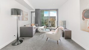 A modern living room with a grey sofa, a white armchair, a round coffee table, and a floor lamp. There is a balcony with a glass door and a view of trees. - lovely ap in  heart of chatswood (Chatswood)