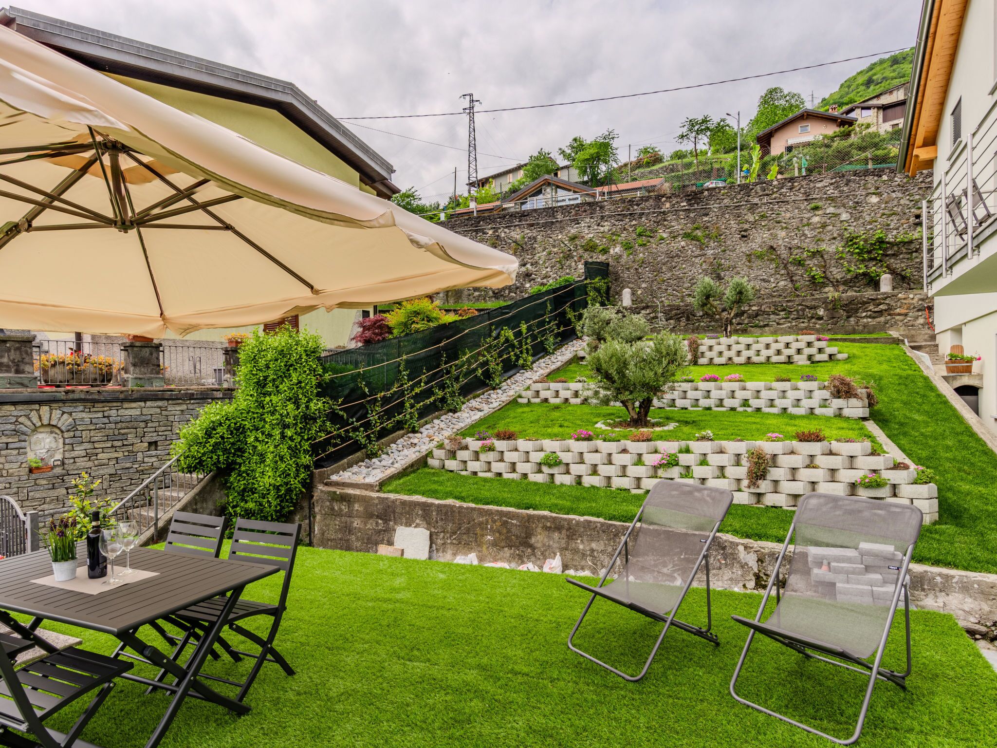 Een terras met een tafel en stoelen, een kleine tuin met een stenen muur en uitzicht op een heuvel.