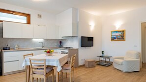 A compact kitchen with white cabinets, a dining table with chairs, a TV mounted on the wall, and a small seating area with a chair and table. - Apartment Syria by Interhome (Domaso)