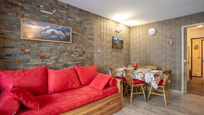 Un salon chaleureux avec un canapé rouge, une table à manger en bois et des chaises. Un tableau représentant une montagne orne le mur, et une horloge est visible sur le mur.
