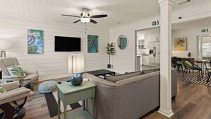Un salon moderne avec un canapé gris, une table d’appoint verte et un ventilateur de plafond. Un téléviseur à écran plat est fixé au mur, et un tableau est accroché au-dessus.