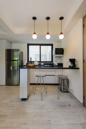 A modern kitchen with a stainless steel refrigerator, a black microwave, and a coffee maker. There are two bar stools and a window above the sink.