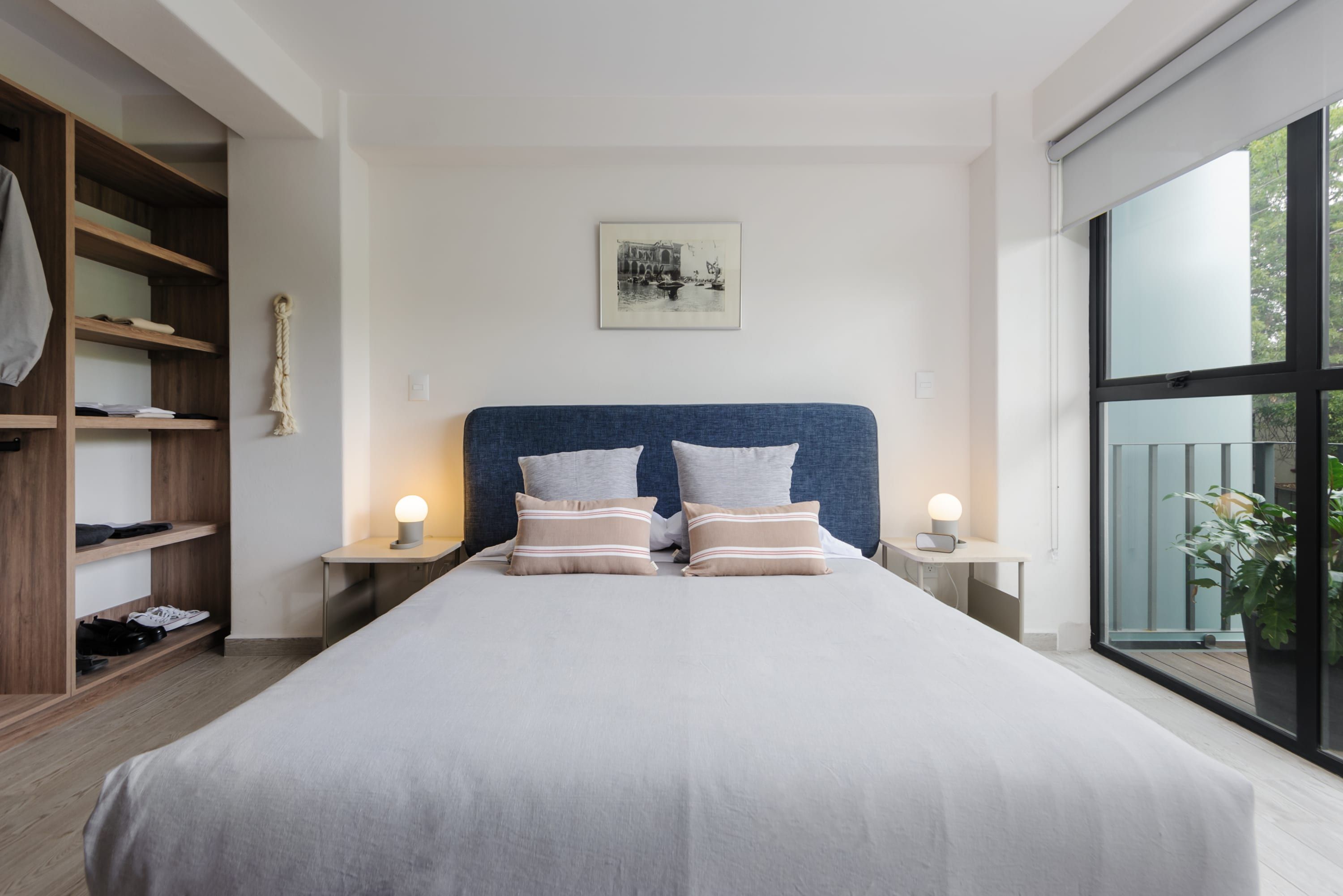 A modern bedroom with a large bed, wooden shelving, a framed picture, bedside lamps, and a large window.